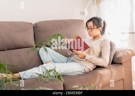 Asiatische Frau liest ein Buch zu Hause. Mädchen auf der Couch entspannen genießen ein altes Buch drinnen während Coronavirus Lockdown. Frau konzentriert lesen. Stockfoto