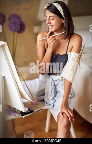 Junge Malerin schaut ihr Gemälde an, lächelnd, zufrieden Stockfoto