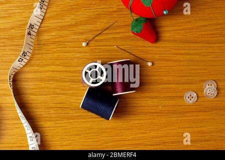 Verschiedene Nähzubehör sitzen auf einem hölzernen Hintergrund. Im Bild kann man ein Nadelkissen mit eingestockten Stiften, Faden, verschiedene Bettwäsche und ein Klebeband sehen Stockfoto