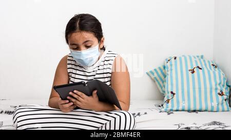 Junge asiatische Mädchen tragen Schutzmaske auf ihrem Bett sitzen mit einem Tablet online zu studieren - Corona Virus Home Schooling Konzept Bild mit Kopie Stockfoto