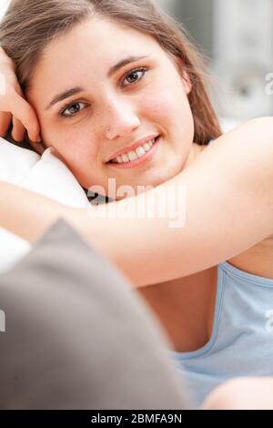 Attraktive, freundliche junge Frau, die sich auf einem Sofa in einem Nahaufnahme-Porträt entspannt, während sie sich gegen die Kissen entspannt und lächelnd die Kamera anlächelt Stockfoto