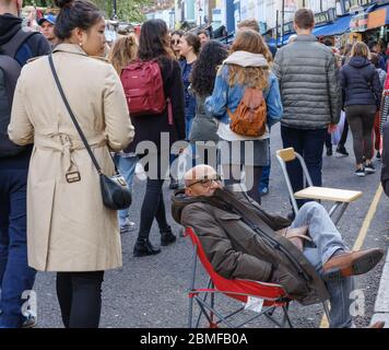 Menschenmenge von Käufern auf dem Portobello Market mit einem verschlafenen Mann im Stuhl, der seinen Laden beobachtet. Kensington & Chelsea, Portobello Road, Notting Hill, London, Großbritannien Stockfoto