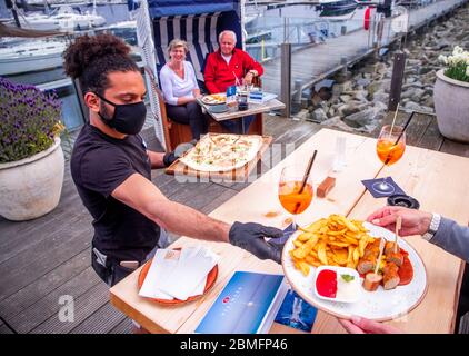 09. Mai 2020, Mecklenburg-Vorpommern, Kühlungsborn: Ein Kellner bedient seine Gäste im Restaurant Vielmeer auf der Terrasse am Seemannshafen mit Mundschutz und Handschuhen. Die Restaurants in Mecklenburg-Vorpommern können Gäste aufgrund der Corona-Schutzmaßnahmen wieder unter strengen Hygienebedingungen und weniger Sitzplätzen bedienen. Seit mehreren Wochen sind Restaurants in ganz Deutschland wegen des Corona-Virus geschlossen. Foto: Jens Büttner/dpa-Zentralbild/dpa Stockfoto