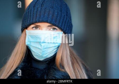 Junge Frau trägt blaue Einweg-Virus Gesicht Mund Nase Maske und warme Mütze Hut, Nahaufnahme Porträt, verschwommener Hintergrund. Coronavirus covid-19 outbrea Stockfoto