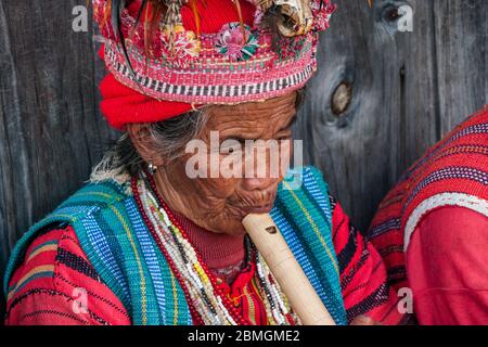 Banaue, Philippinen - Februar 2012: Ältere Frau in traditioneller Kleidung sitzt vor einer Holzwand und spielt Flöte Stockfoto