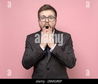 Überraschender Mann mit Brille und Anzug schaut mit großen Augen auf die Kamera und bedeckt seinen offenen Mund mit den Händen. Stockfoto