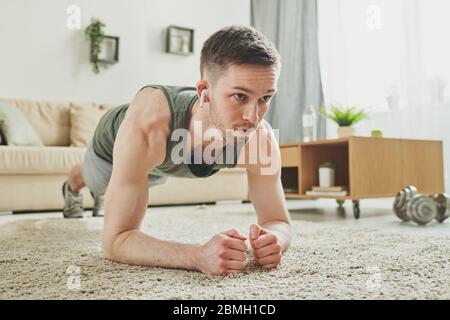 Junger aktiver Mann mit Ohrhörern, der während der Quarantänezeit zu Hause im Wohnzimmer eine Plankenübung auf dem Teppich macht Stockfoto