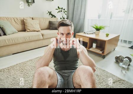 Junge ernsthafte schweißtreibende Athlet macht Sit-ups vor der Kamera und schaut Sie an, während Sie auf dem Teppich in der häuslichen Umgebung sitzen Stockfoto