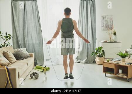 Rückansicht des jungen Mannes im Aktivspringen mit Springseil während er vor dem Fenster des Wohnzimmers während des Trainings steht Stockfoto