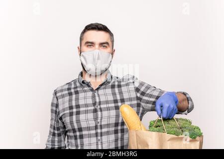 Junger Mann im karierten Hemd, Schutzmaske und Handschuhe halten Paperbag mit frischem Brot, Brokkoli und andere Lebensmittel isoliert Stockfoto