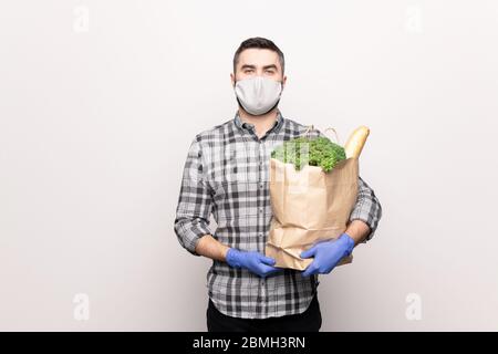 Ernst junger Mann in Schutzmaske und Handschuhe halten Papierpaket mit frischen Lebensmitteln, während sie zu Ihnen nach Hause liefern Stockfoto