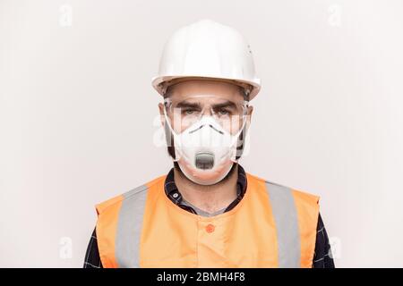 Seriöse männliche Techniker in orange Arbeitskleidung, Schutzbrille und Atemschutzmaske Blick auf Sie, während auf weißem Hintergrund stehen Stockfoto