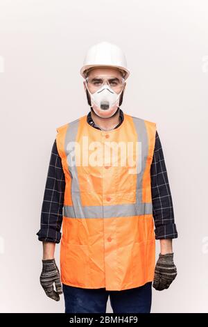 Junger ernsthafter Techniker in orangefarbener Arbeitskleidung, Schutzhandschuhen, Brillen und Atemschutzmaske, die vor der Kamera auf weißem Hintergrund stehen Stockfoto