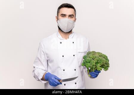 Junger männlicher Koch in Schutzmaske, Handschuhe und weiße Uniform hält scharfe Messer und frischen Brokkoli, während gehen, Gemüsesuppe zu kochen Stockfoto
