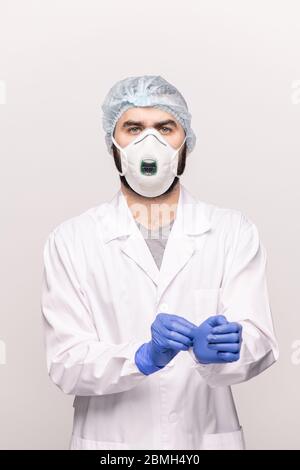 Junger Chirurg in Weißkoat, medizinische Kappe, Atemschutzmaske und Brille, die schützende medizinische Handschuhe auf seine Hände vor der Operation Stockfoto