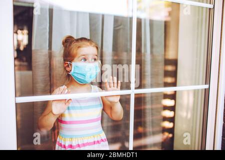 Kleines Mädchen trägt eine medizinische Maske in Quarantäne, das hinter einem großen Fenster mit weit geöffneten Augen bleibt. Quarantänekonzept des Coronavirus. Stockfoto