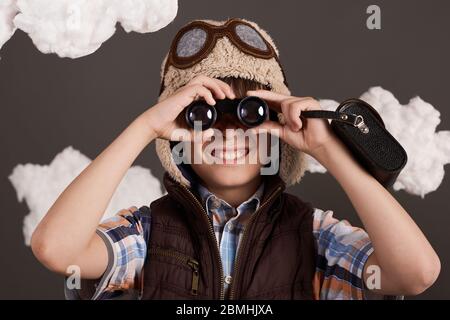 Ein Junge spielt mit Ferngläsern und träumt davon, Pilot zu werden, gekleidet in eine Retro-Jacke und Helm mit Brille, Wolken aus Watte, grauer Rücken Stockfoto