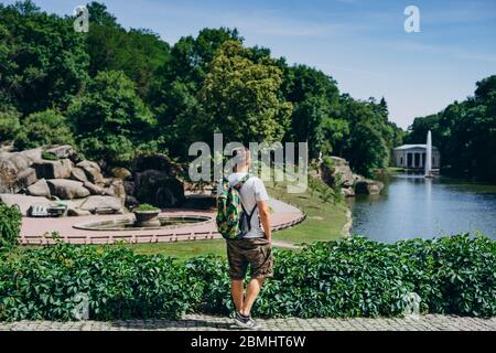 Sofia Park, Ukraine. Mann mit Rucksack in einem Landschaftsgarten im Sommer. Der Mensch drehte den Rücken auf dem Hintergrund des Sees mit einem Brunnen. Stockfoto