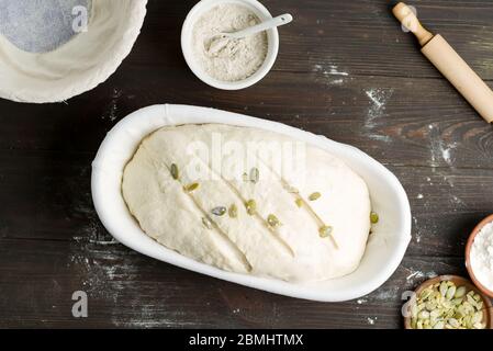 Frisch zubereitetes Laib selbstgemachten weißen gesunden Brot mit Kürbiskern in einem Teigkorb ist bereit zum Backen auf einem dunklen Holzhintergrund, Kopierraum. Stockfoto