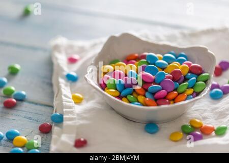 Smarties in eine weiße Porzellanschale legen und auf einer weißen Umkleide und einem Tisch verstreut. Stockfoto