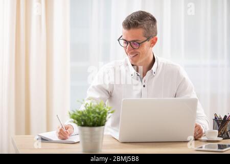 Geschäftsmann sitzt an seinem Schreibtisch hinter seinem Notizbuch und schreibt sich Notizen, lächelnd. Stockfoto