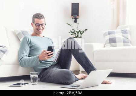 Lächelnder Mann SMS auf seinem Handy während seiner Bürozeiten zu Hause, auf einem Wohnzimmer Boden sitzen. Stockfoto