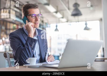Ein Geschäftsmann ruft mit einem Handy an und schaut in das Notebook, um im Restaurant einen Kaffee zu trinken. Stockfoto