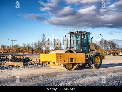 Auffällige gelbe Straßenwalze mit geschlossener, klimatisch gesteuerter Kabine steht auf nicht fertig neuer Straße, Steine, blauer Himmel, Wolken, Vorderansicht rechts. Stockfoto