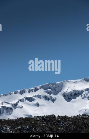 Nahaufnahme des schneebedeckten Bergrückens mit blauem Himmel und Bäumen darunter Stockfoto