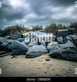 Quadratische lange Aufnahme eines dramatischen bewölkten Himmels mit vielen riesigen Felsen an einem Sandstrand. Stockfoto