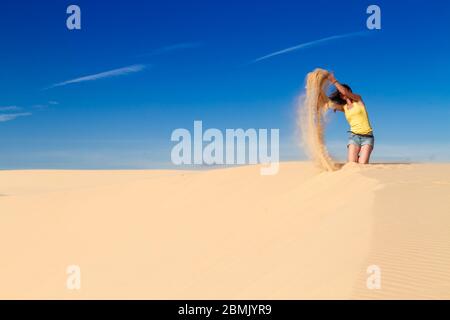 Frau sitzt auf dem Sand auf einem Bergrücken in der Wüste, wirft Sand in die Luft. Stockfoto