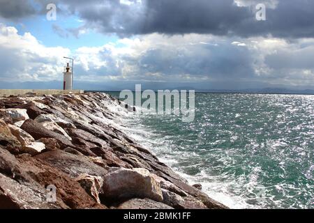 Ein Hafen aus Felsen und ein weißer Leuchtturm. Welliges Meer und wolkiges windiges Wetter. Stockfoto