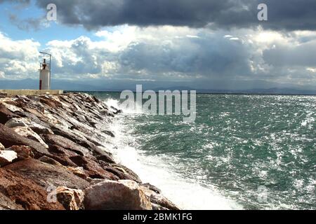 Ein Hafen aus Felsen und ein weißer Leuchtturm. Welliges Meer und wolkiges windiges Wetter. Stockfoto