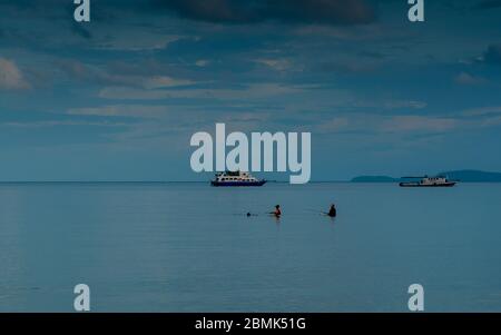 3 Personen stehen in ruhigem Wasser mit Booten im Hintergrund mit Wolken und blauem Himmel im Hintergrund Stockfoto