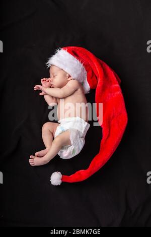 Kleines Baby auf dunklem Hintergrund in einem langen weihnachtsmann Hut, der in der Form eines Halbmonds liegt Stockfoto