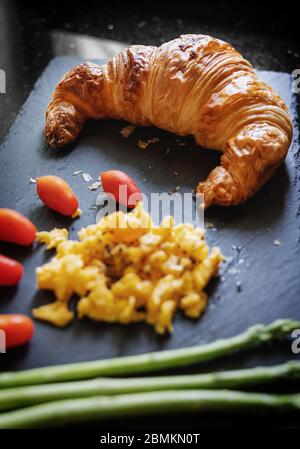 Frischer Croissant mit Rührei, Kirschtomaten und Spargel auf schwarzem Steinteller Stockfoto