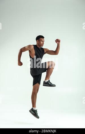 Do it volle Länge des jungen afrikanischen Mann mit muskulösen Körper in Sportbekleidung Springen im Studio vor grauem Hintergrund. Sportkonzept. Motivation. Gesunde Lebensweise Stockfoto