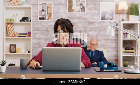 Ältere Altersfrau, die auf dem Laptop einen Schluck Kaffee trinkt. Der alte Mann erholte sich auf dem Sofa im Hintergrund. Stockfoto