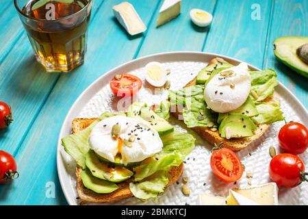 Gesundes Sandwich - pochierte Eier und Avocado auf Toast mit Tomaten auf blauem Holzhintergrund. Stockfoto
