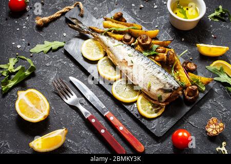 Makrele mit Zitrone, gebackenen Kartoffeln und Pilzen auf schwarzem Steingrund. Stockfoto