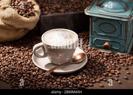 Heiße Kaffeetasse Cappuccino oder Latte, geröstete Bohnen und alte Mühle. Rustikaler Kaffee Hintergrund Stockfoto