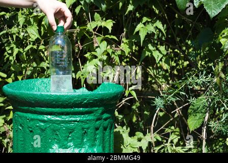 Weibliche Hand wirft eine Plastikflasche in einen kleinen Eisenmüllbehälter in einem grünen Park im Sommer, Nahaufnahme. Naturschutz vor Plastik Stockfoto