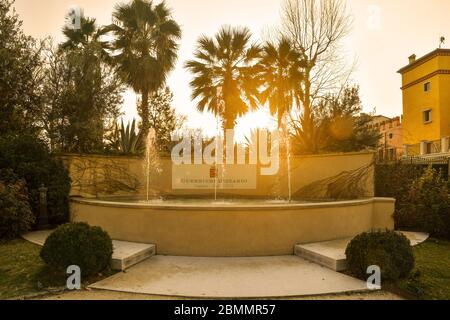 Blick von hinten auf einen Steinbrunnen im Park der Villa Guerrieri am Ufer des Gardasees mit Palmen bei Sonnenuntergang, Bardolino, Verona, Veneto, Italien Stockfoto