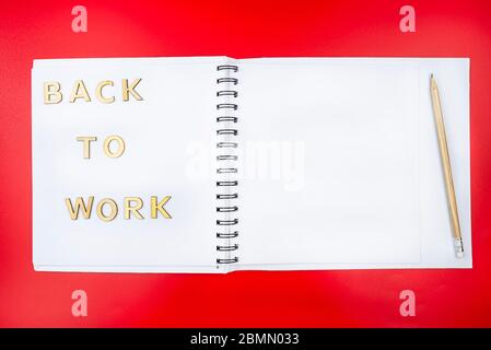 Die Inschrift "Back to work" aus Holzbuchstaben, die auf einem offenen Notizbuch mit weißen Seiten und einem Bleistift liegen, isoliert auf rotem Hintergrund. Stockfoto