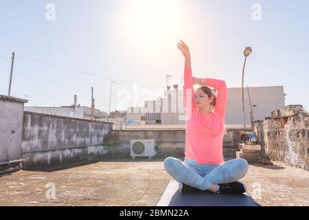 Das junge Mädchen dehnt sich während der Sperrung des Coronavirus auf der Matte auf dem Dach ihres Hauses aus. Frauen Yoga-Training für gesund zu bleiben. Selbstversorgung Stockfoto