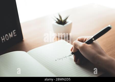 Frau Hand mit Bleistift Schreiben Bleiben Sie sicher auf dem Notebook zu Hause Schreibtisch. Arbeit von zu Hause und Quarantäne-Konzept auf Epidemien Ausbruch. Stockfoto