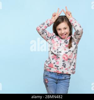 Studioaufnahme eines ausdrucksstarken niedlichen Kindes, das die Finger wie Häschen oder Hörner hinter dem Kopf hält, die Kamera anschaut und lächelt. Lifestyle und schöne p Stockfoto