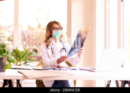 Aufnahme einer Ärztin, die Gesichtsmaske trägt, während sie im Zimmer des Arztes sitzt und das Röntgenbild überprüft. Stockfoto