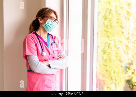 Porträtaufnahme einer überarbeiteten Krankenschwester, die eine Gesichtsmaske trägt, während sie am Fenster steht und nach draußen schaut. Stockfoto