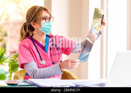 Aufnahme einer Ärztin, die Gesichtsmaske trägt, während sie im Zimmer des Arztes sitzt und das Röntgenbild überprüft. Stockfoto